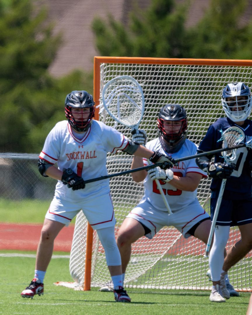 Three lacrosse players in white and dark uniforms defending the goal on a grassy field