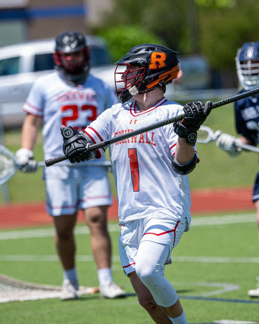 Lacrosse player in white uniform with orange helmet cradling a stick on a field, teammates blurred behind.