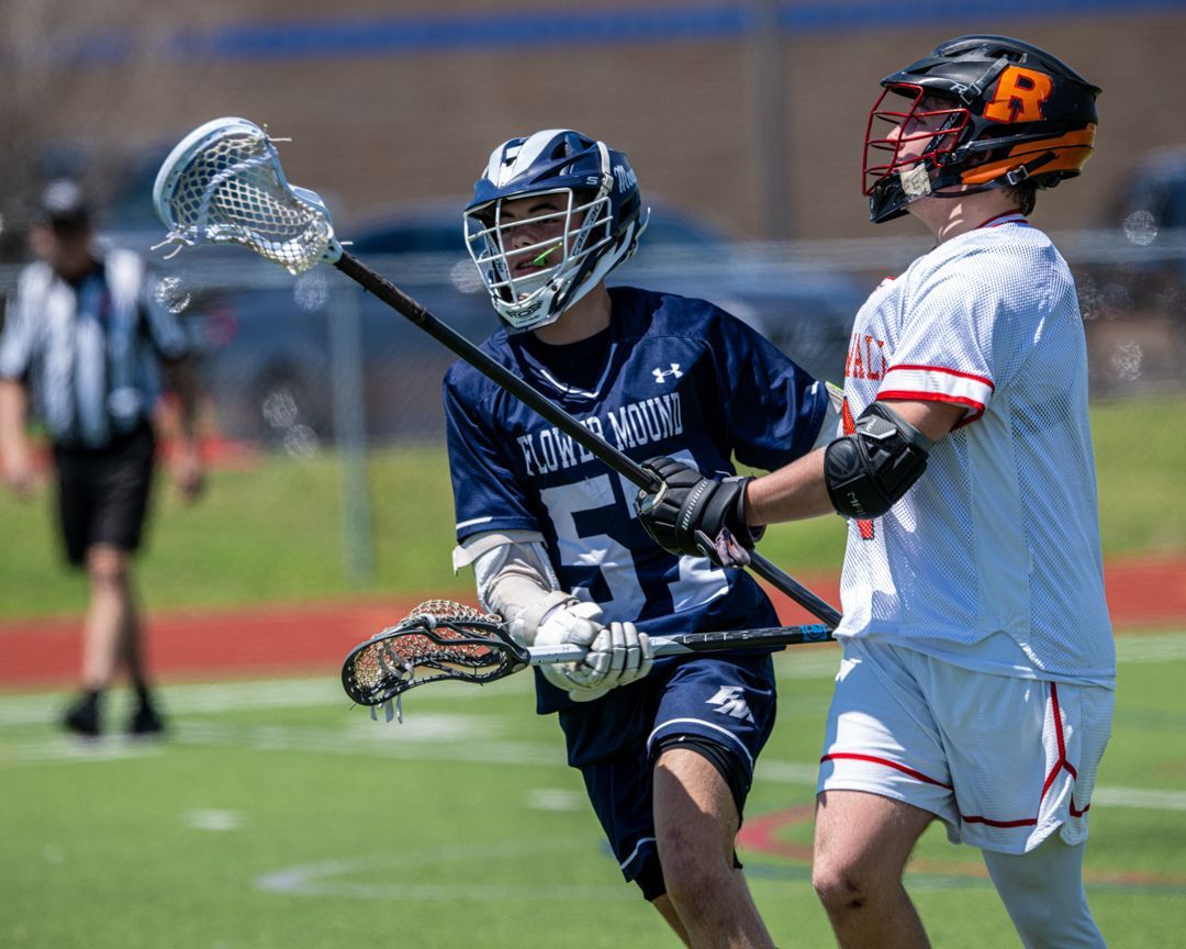 Two lacrosse players in navy and white uniforms compete for the ball on a field.