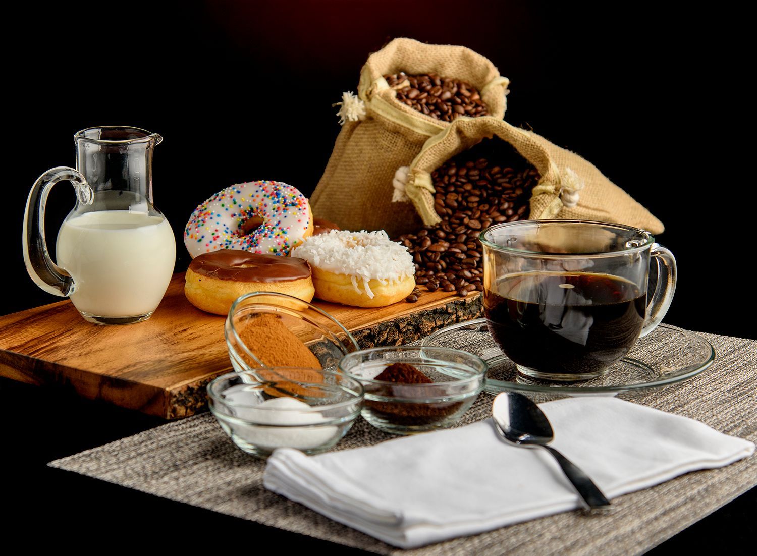 Coffee, milk, donuts, and sugar in sacks on a wooden board with napkins and spoon on a black background