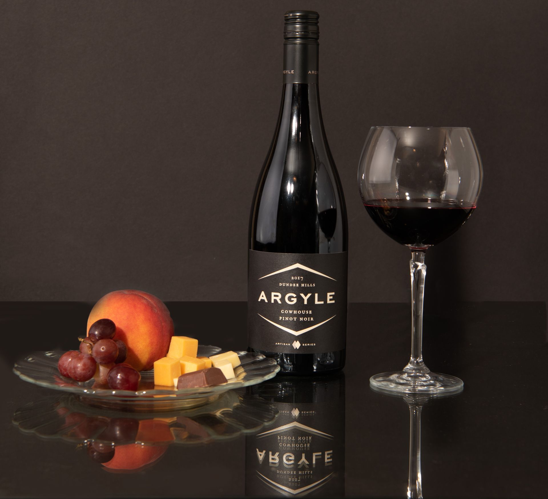 Wine bottle and glass beside a plate of fruit and cheese on a dark reflective surface