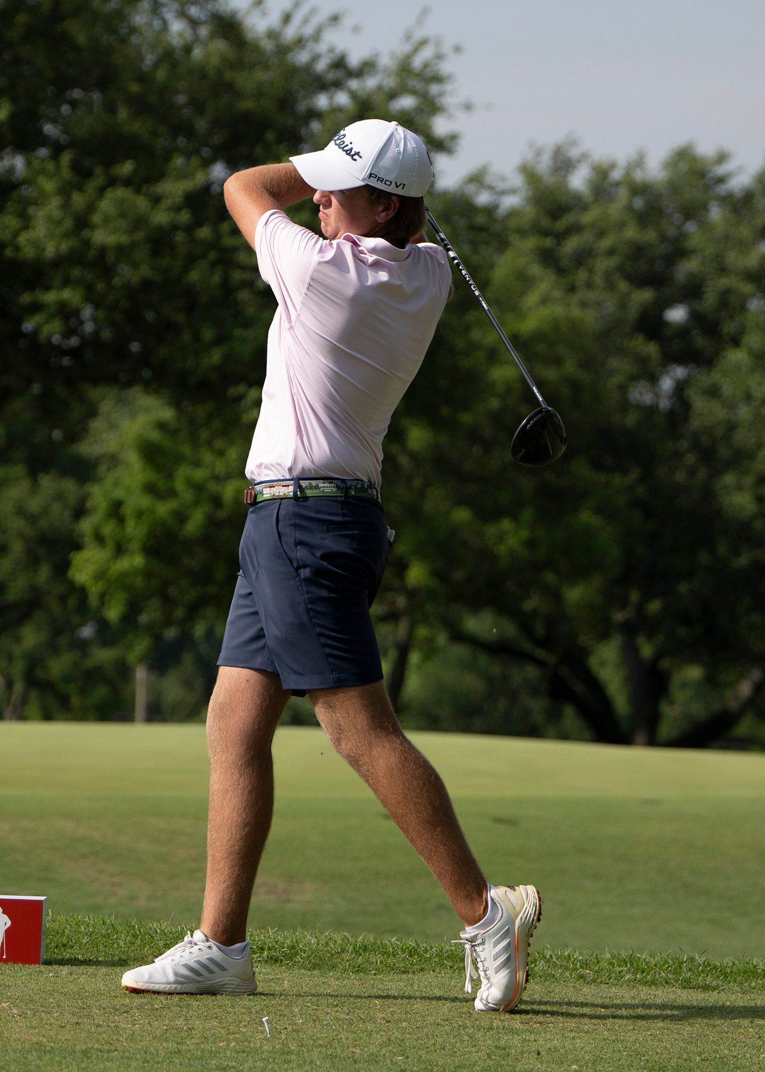 Golfer in white shirt and navy shorts following through on a golf swing on a green course
