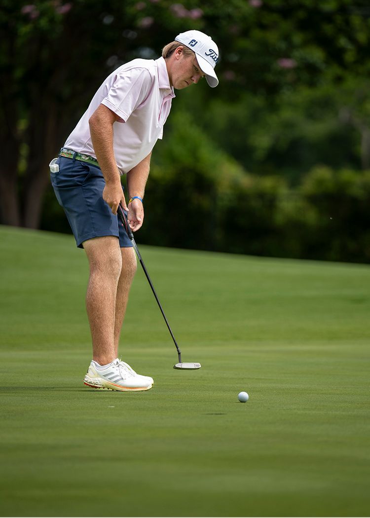 Golfer putting on a green, wearing a white cap and shirt with blue shorts.