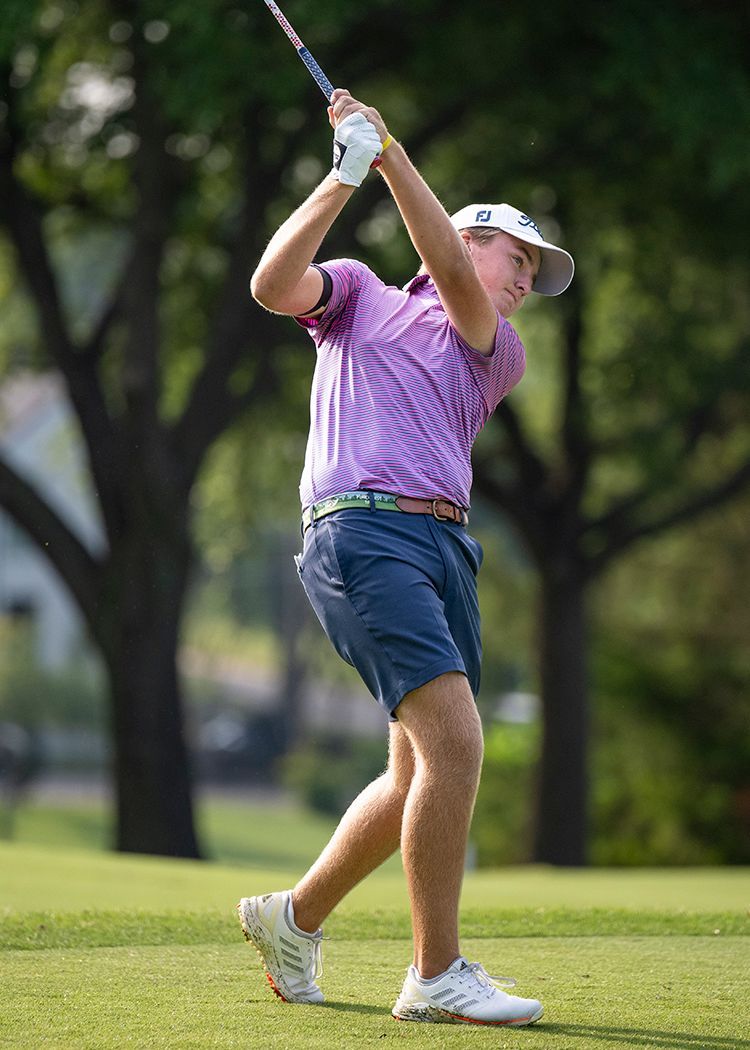 Golfer mid-swing on a grassy course, wearing a purple shirt and blue shorts