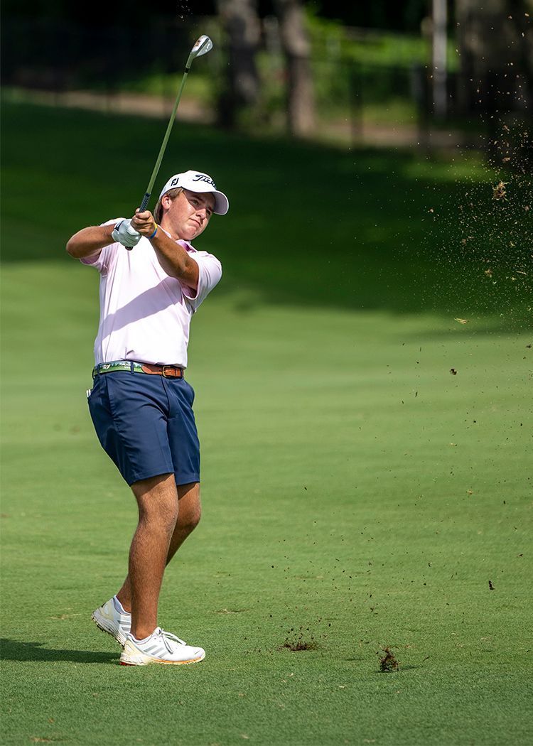 Golfer in white shirt and blue shorts swinging a club on a green course, sand flying nearby