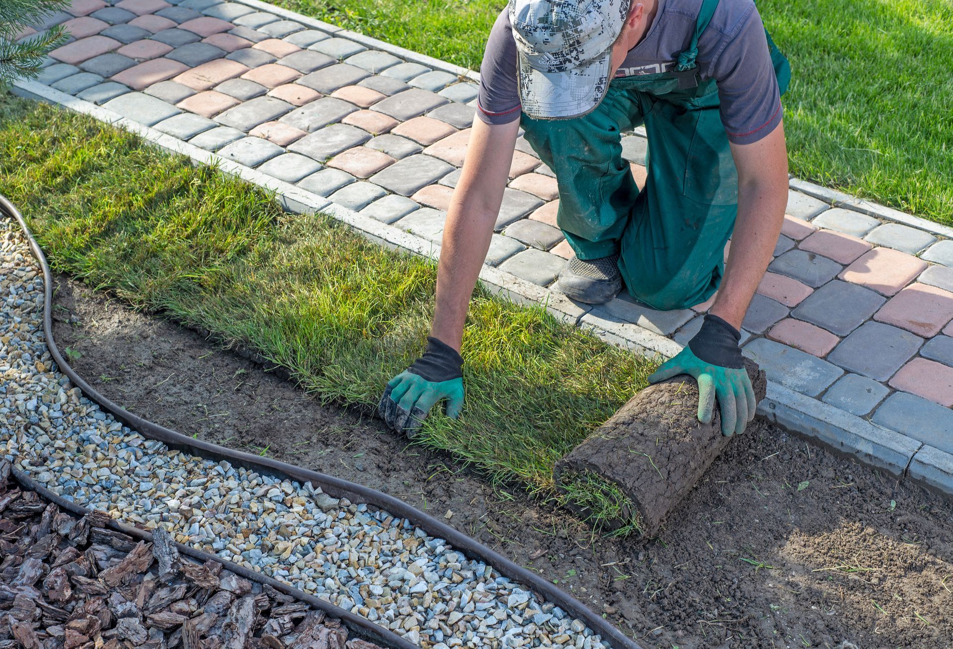 Landscaper unrolling fresh sod beside a paver walkway and gravel bed