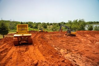 Two construction vehicles on a red dirt road, one in the foreground and an excavator at right.