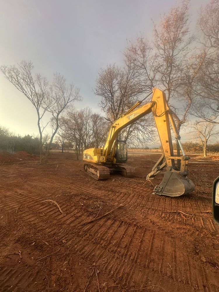 Yellow excavator on a dirt lot under bare trees at dusk