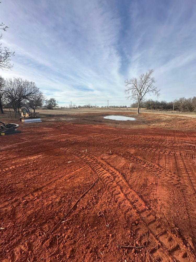 Red dirt field with tire tracks, shallow puddles, bare trees, and a cloudy blue sky