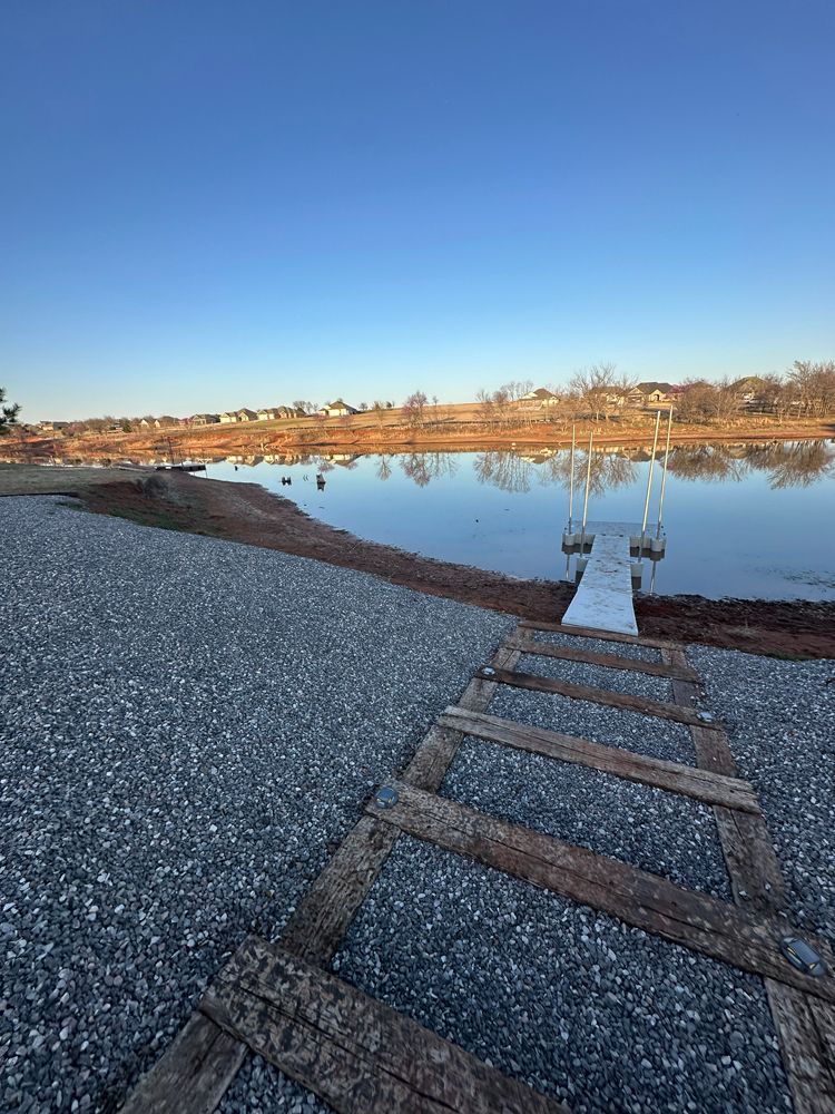 Wooden dock extending into a calm pond beside a gravel shore under a clear blue sky