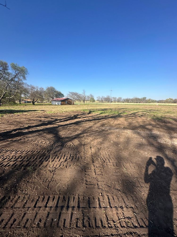 Dirt field under clear blue sky with tire tracks, trees, and a small barn in the distance