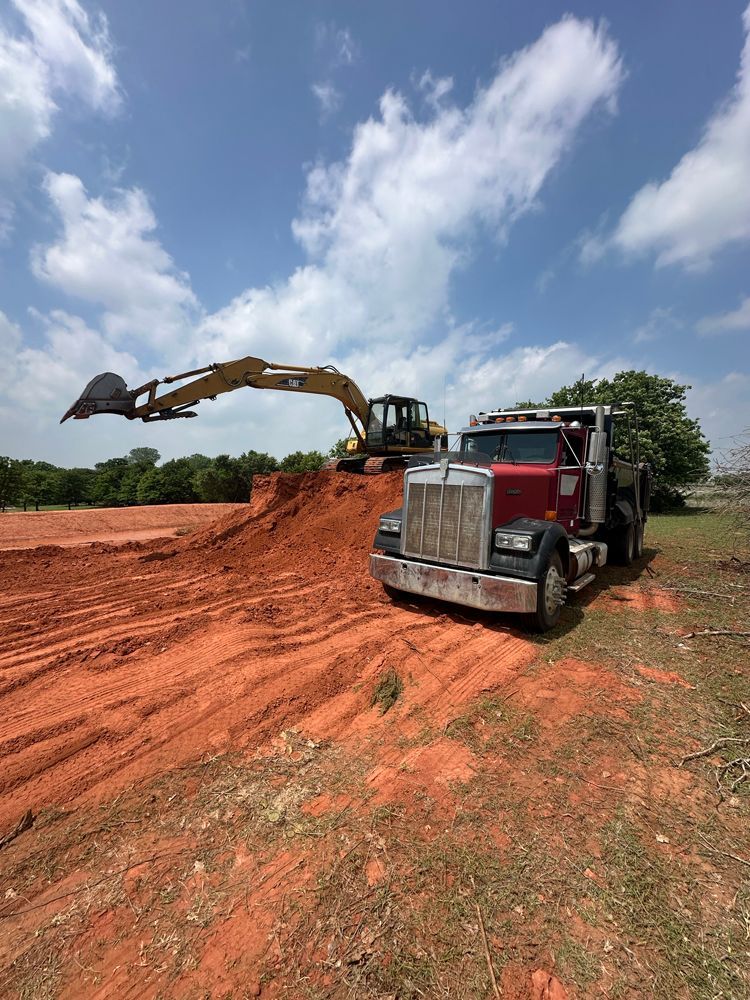 Excavator loading red dirt into a dump truck at a construction site under a cloudy sky