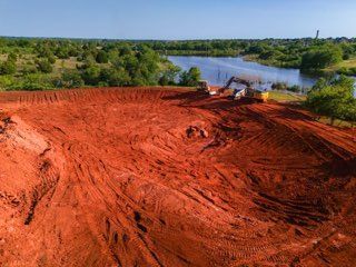 Red dirt construction site beside a river, with heavy machinery and green trees under a blue sky