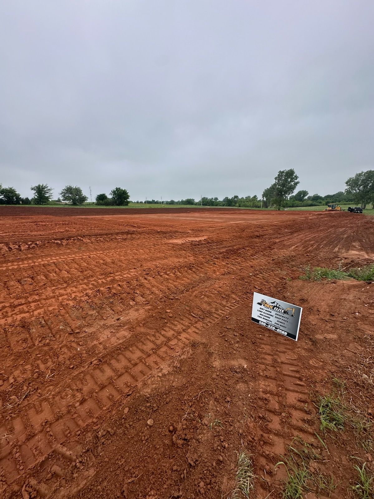 Red dirt field under cloudy sky with a small sign in the foreground and trees along the horizon
