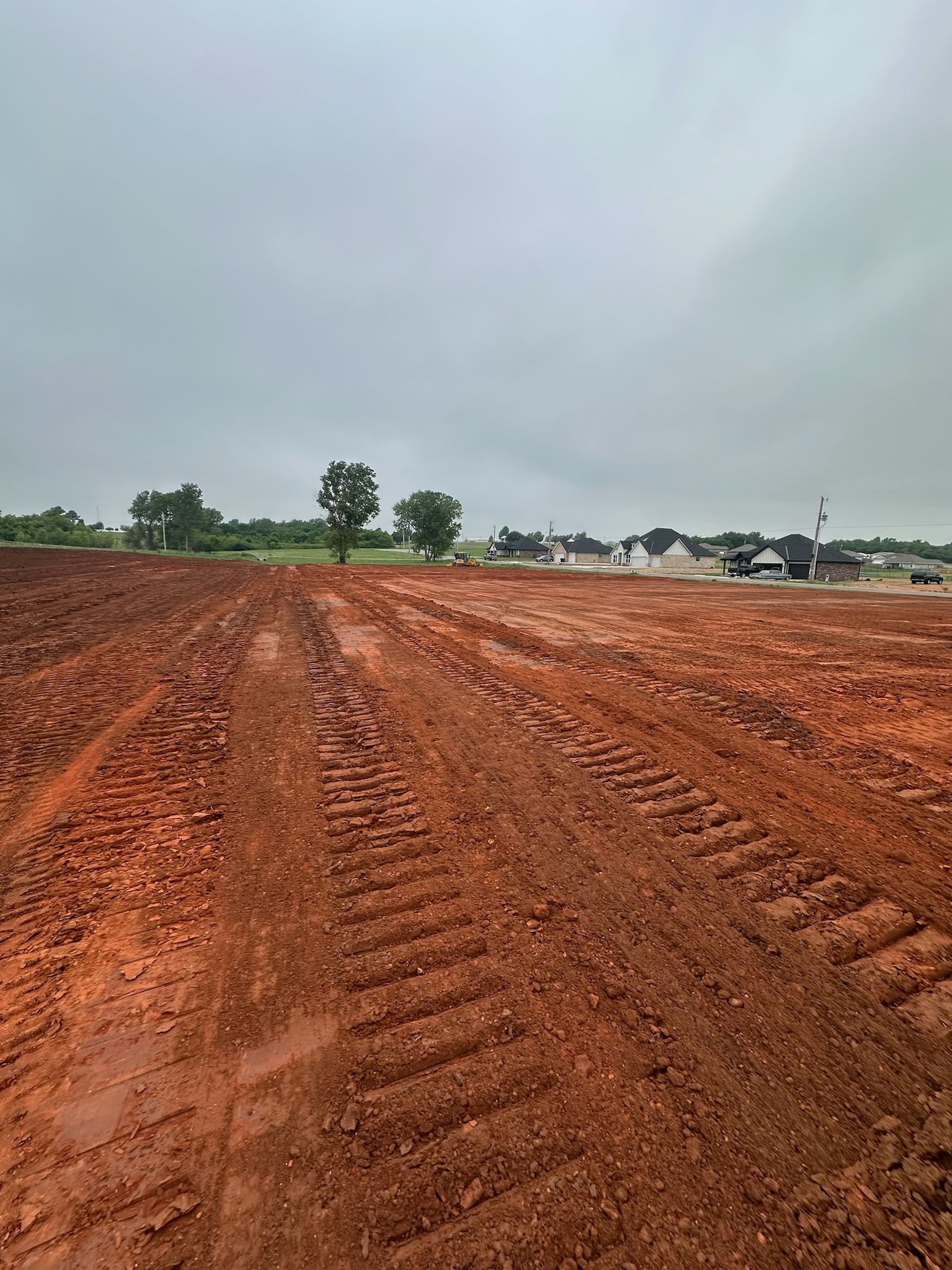 Red dirt field under cloudy sky with tire tracks leading toward distant houses and trees.