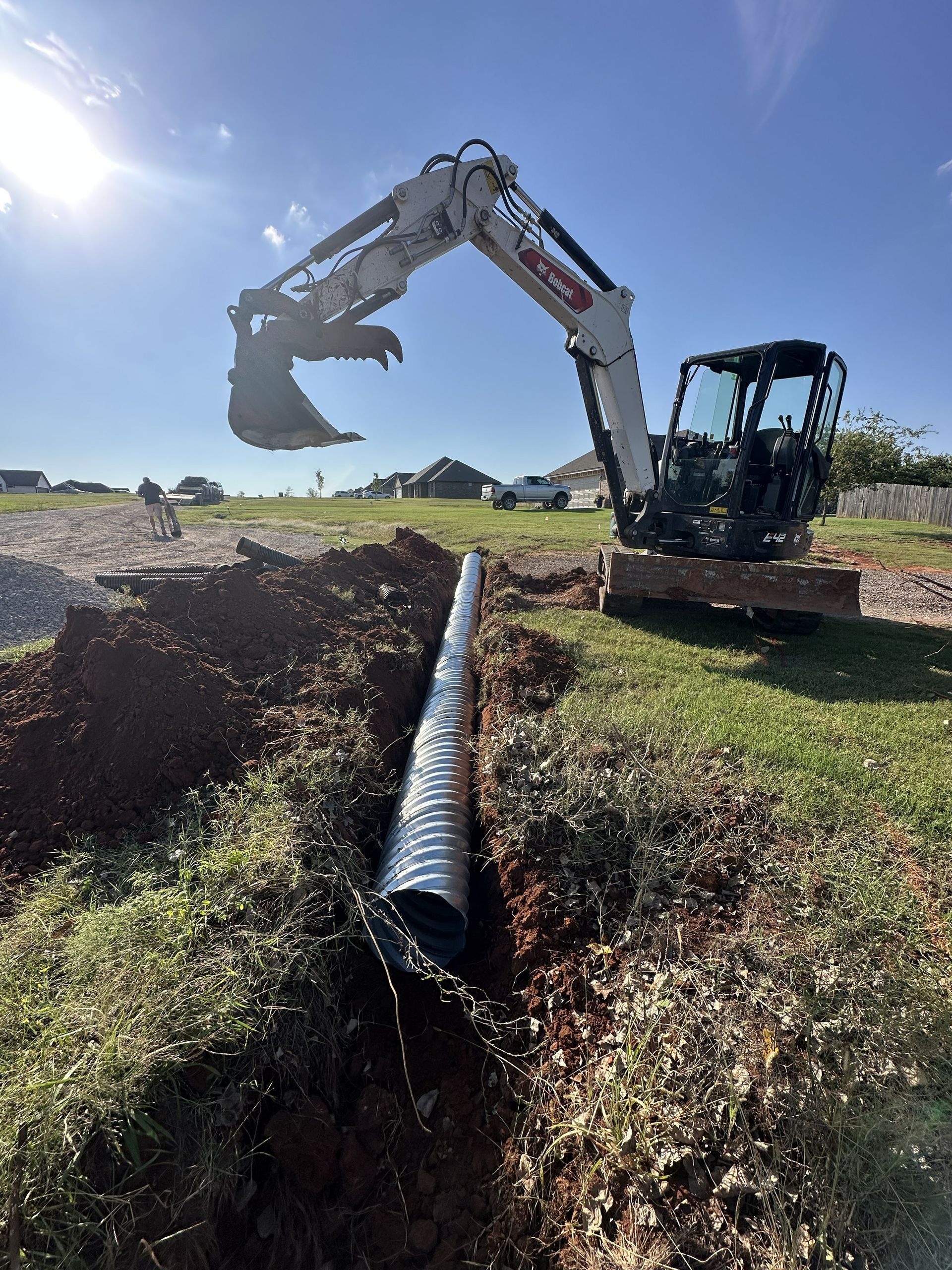 Excavator lifting a large black pipe over a grassy trench under a bright blue sky.