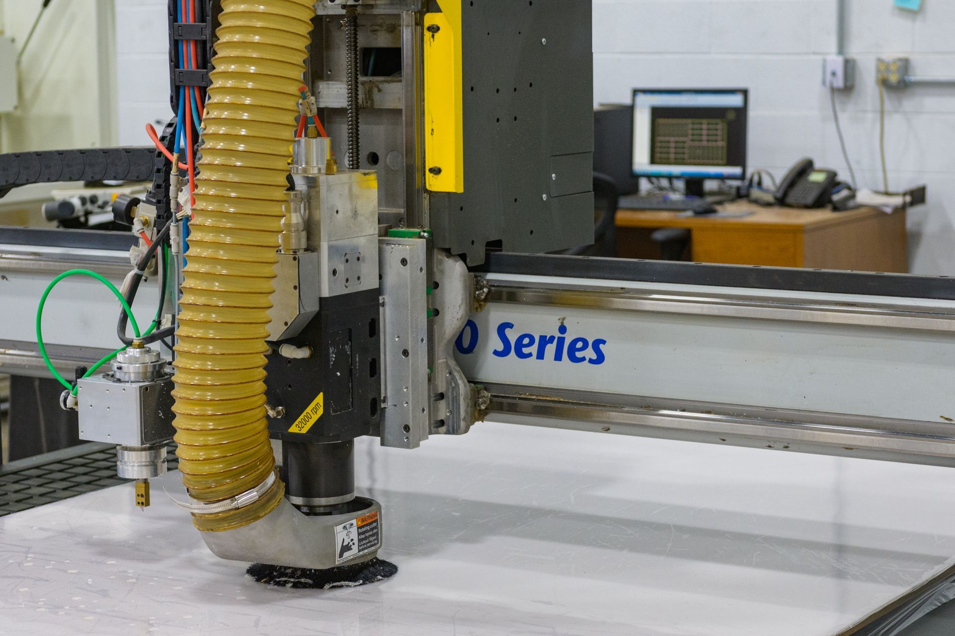 A CNC router cutting a white material sheet in a workshop, with a yellow dust extraction hose attached to the spindle.