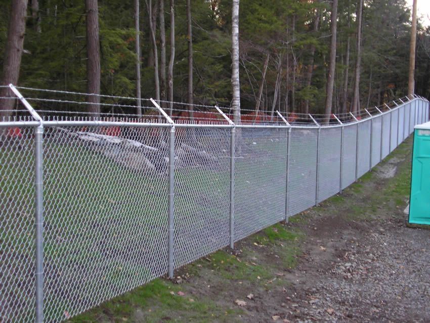 Chain-link fence along a gravel path beside a wooded area, with a green utility box on the right.