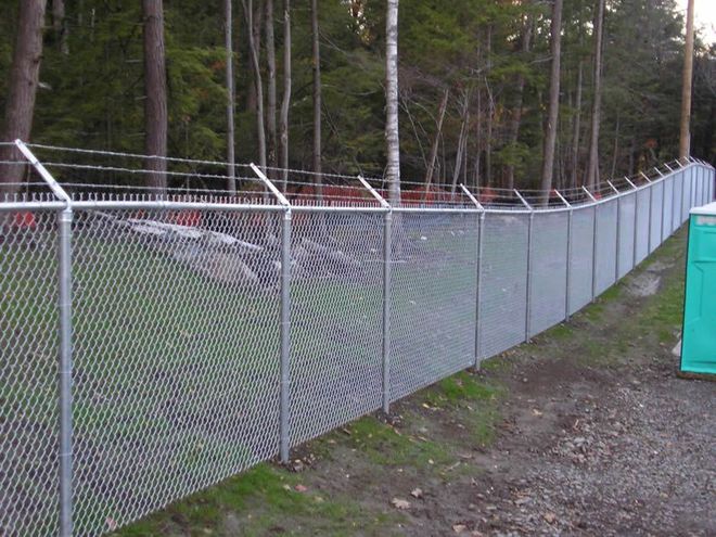 Chain-link fence along a gravel path beside a wooded area, with a green utility box on the right.