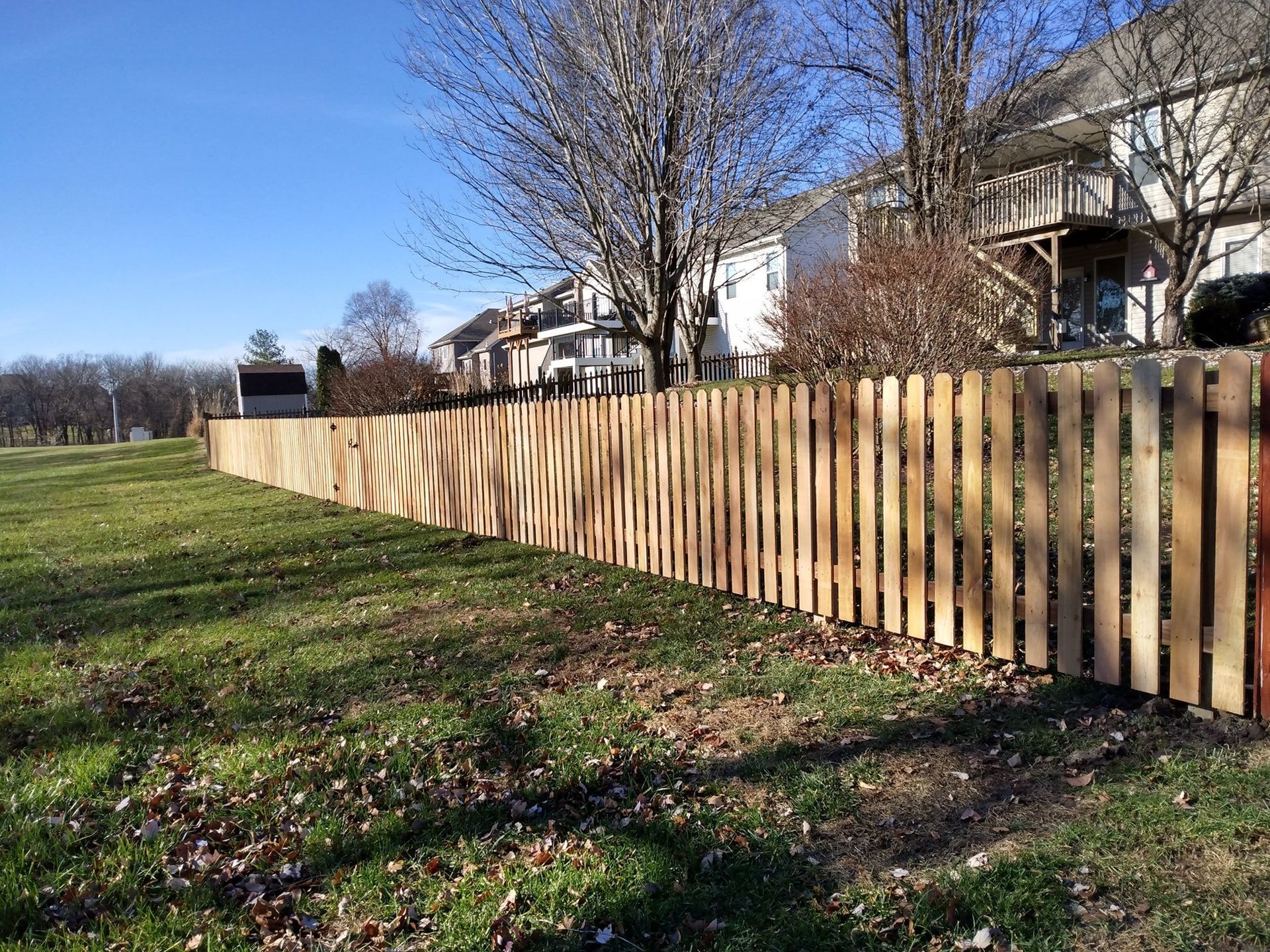 Long wooden fence beside a grassy field under a clear blue sky, with houses in the background