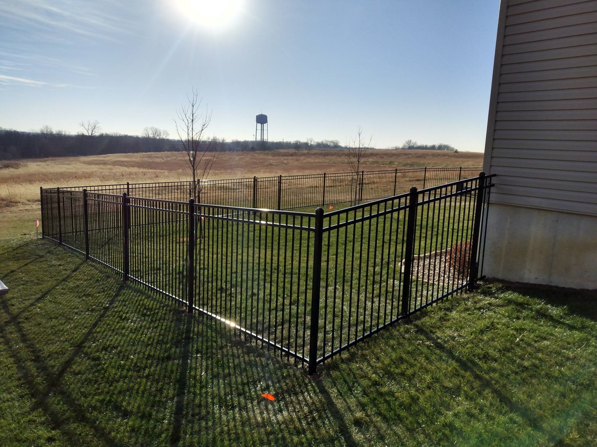 Black metal fence enclosing a grassy yard beside a house, with open field and water tower in the distance.