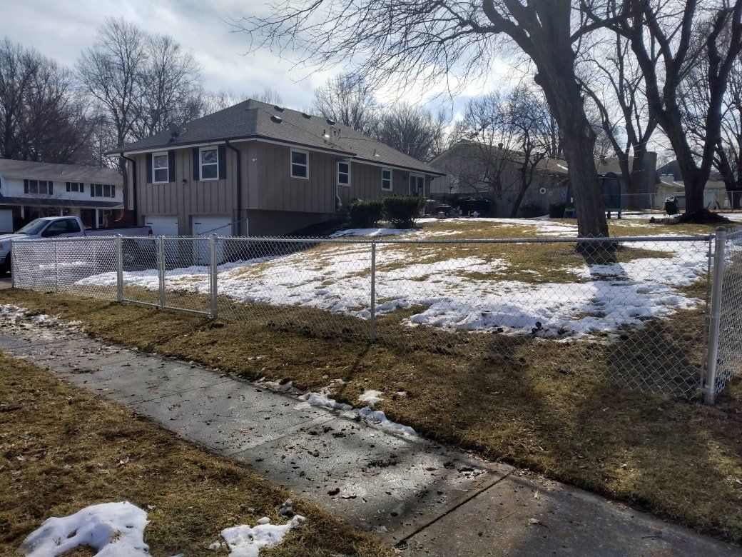 Snowy yard with chain-link fence beside a tan house and leafless trees under a cloudy sky