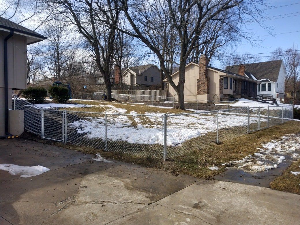 Snowy residential yard with a chain-link fence and houses in the background