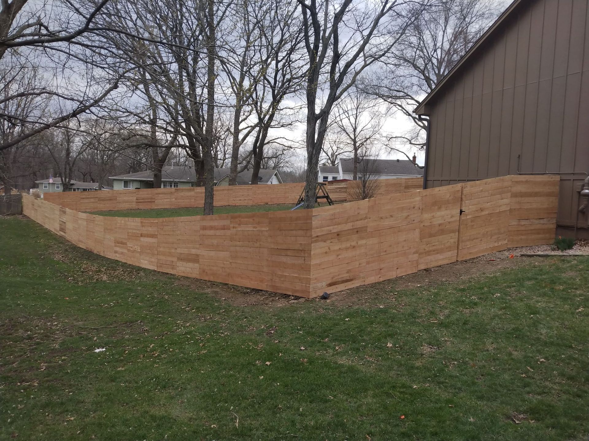 Wooden privacy fence enclosing a backyard beside a house, with leafless trees and grass.