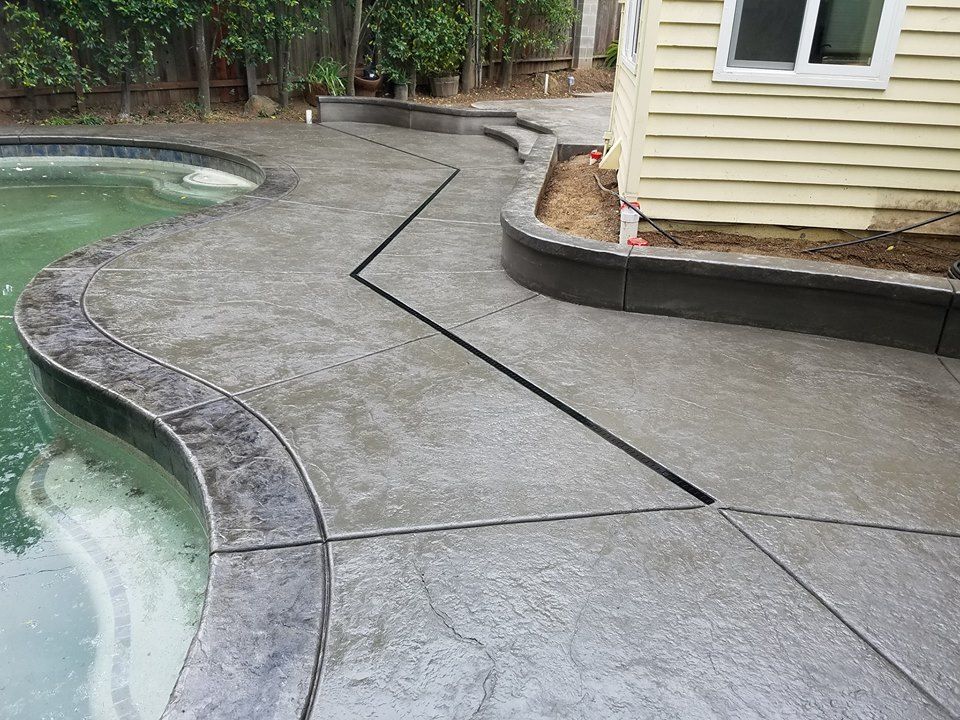 A concrete patio next to a swimming pool with a house in the background.