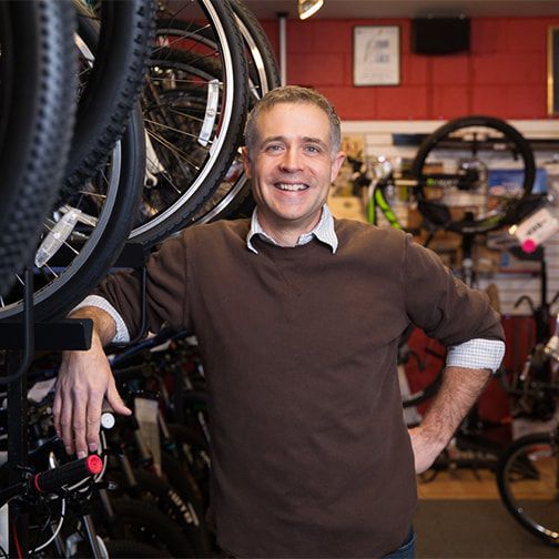 A man in a brown sweater is standing in front of a rack of bicycles.