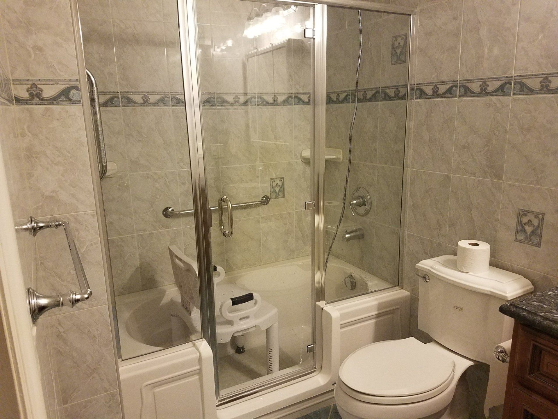 Tiled bathroom with a glass shower enclosure around a step-in tub, toilet, sink, and a paper towel roll on the counter.