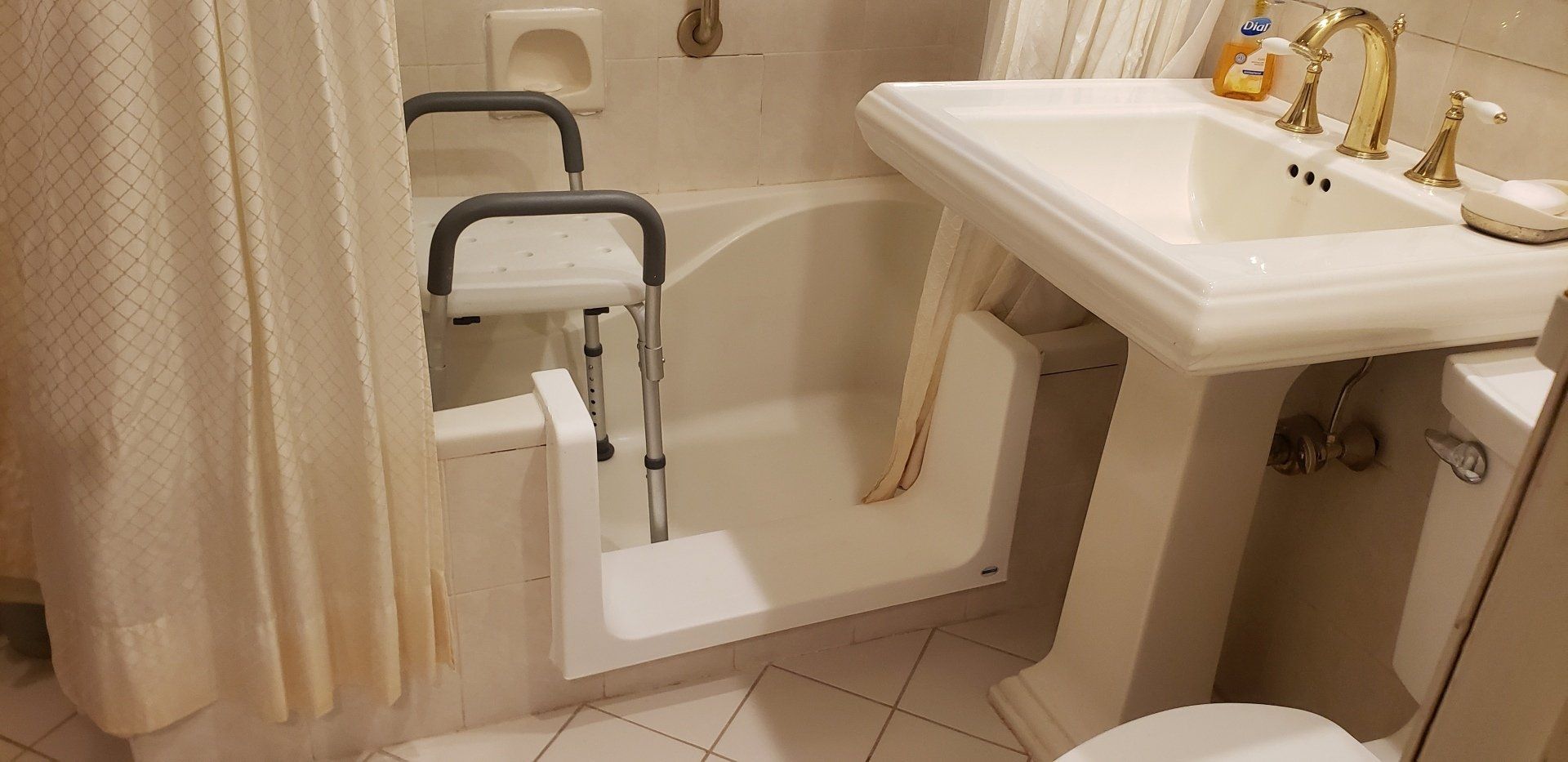 Bathroom with a white pedestal sink, beige tile, and a shower chair beside the step-in bathtub.