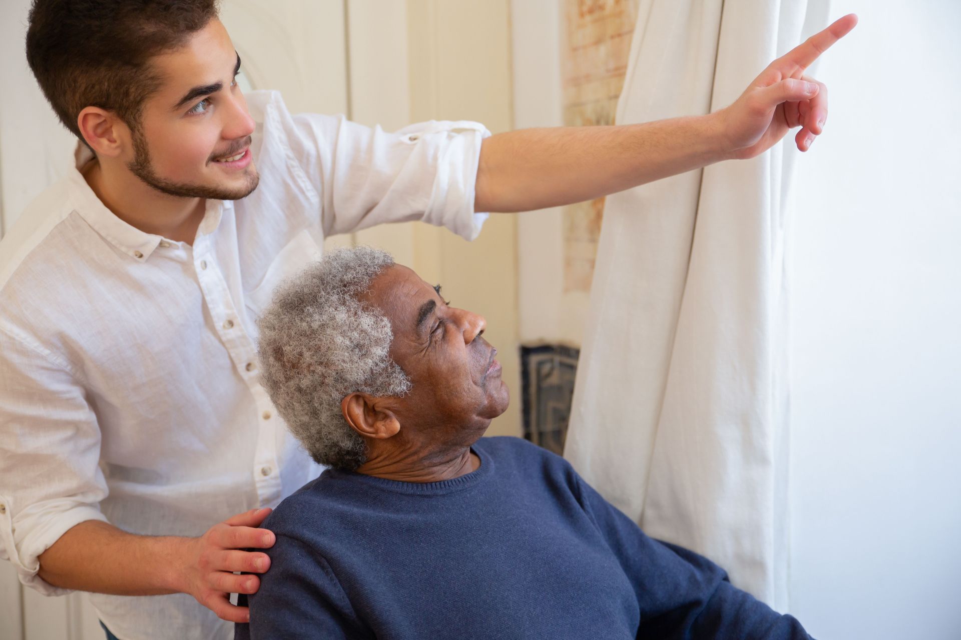 Caregiver points toward a window while supporting an elderly person seated in a bright room