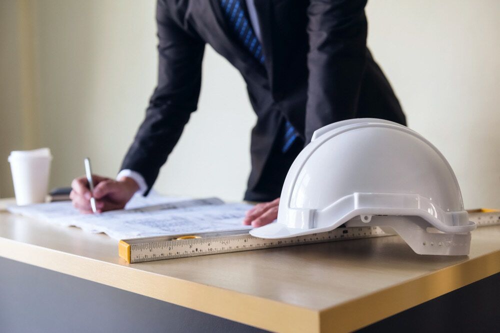 A Man In A Suit And Tie Is Writing On A Blueprint Next To A Hard Hat — GEO – AM Consulting In Lismore, NSW