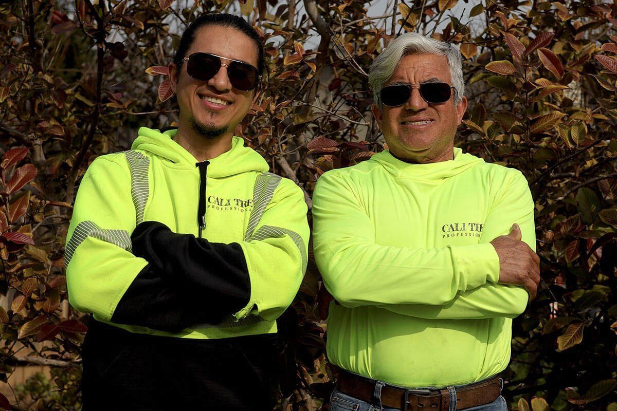 Two people in neon yellow shirts and sunglasses stand with arms crossed outdoors among autumn leaves.