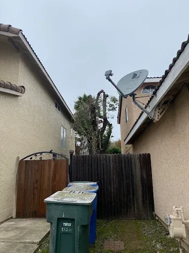 Narrow alley between houses with trash bins, wooden fence, and a satellite dish on the roof.