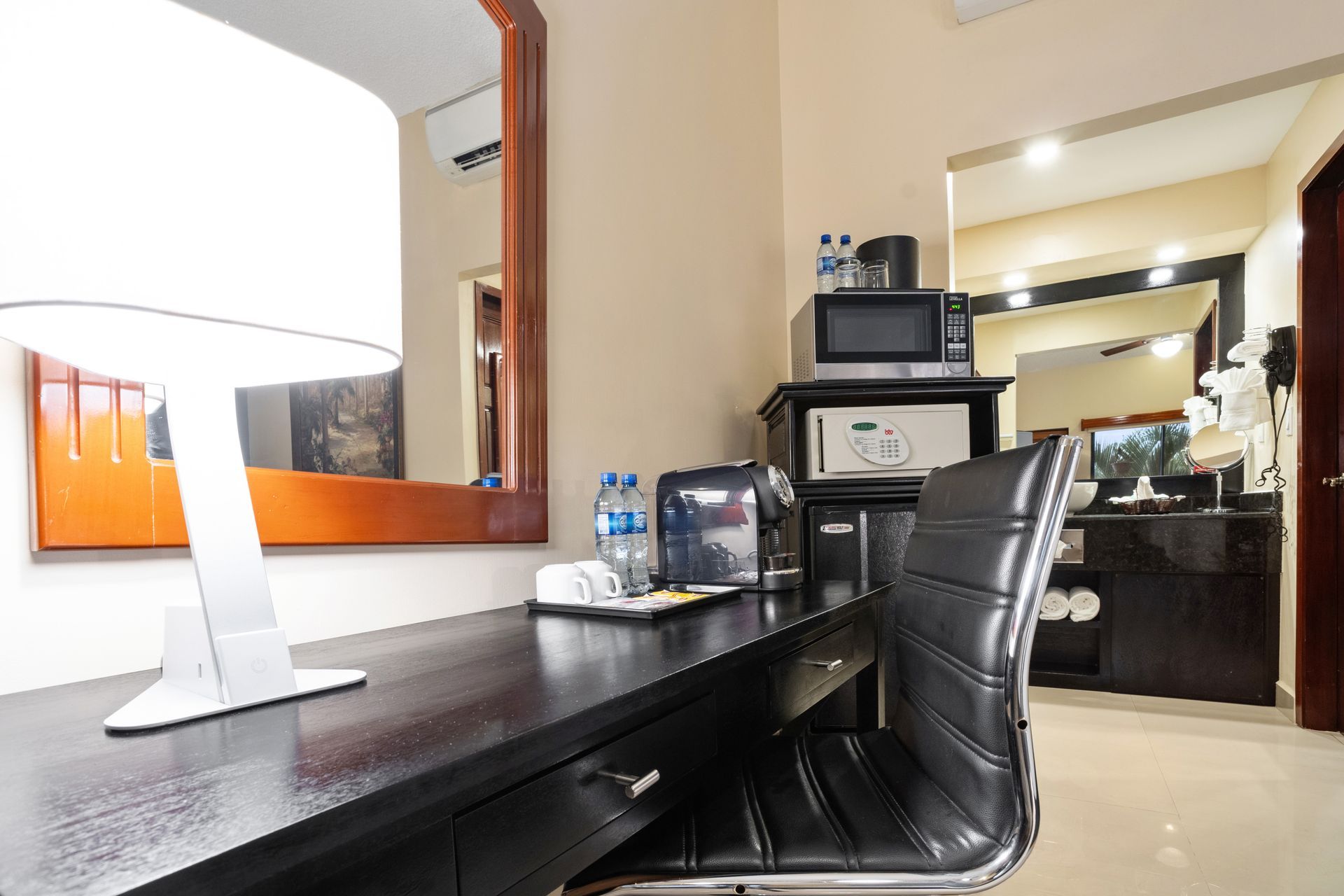 A hotel room with a desk , chair , mirror and microwave.