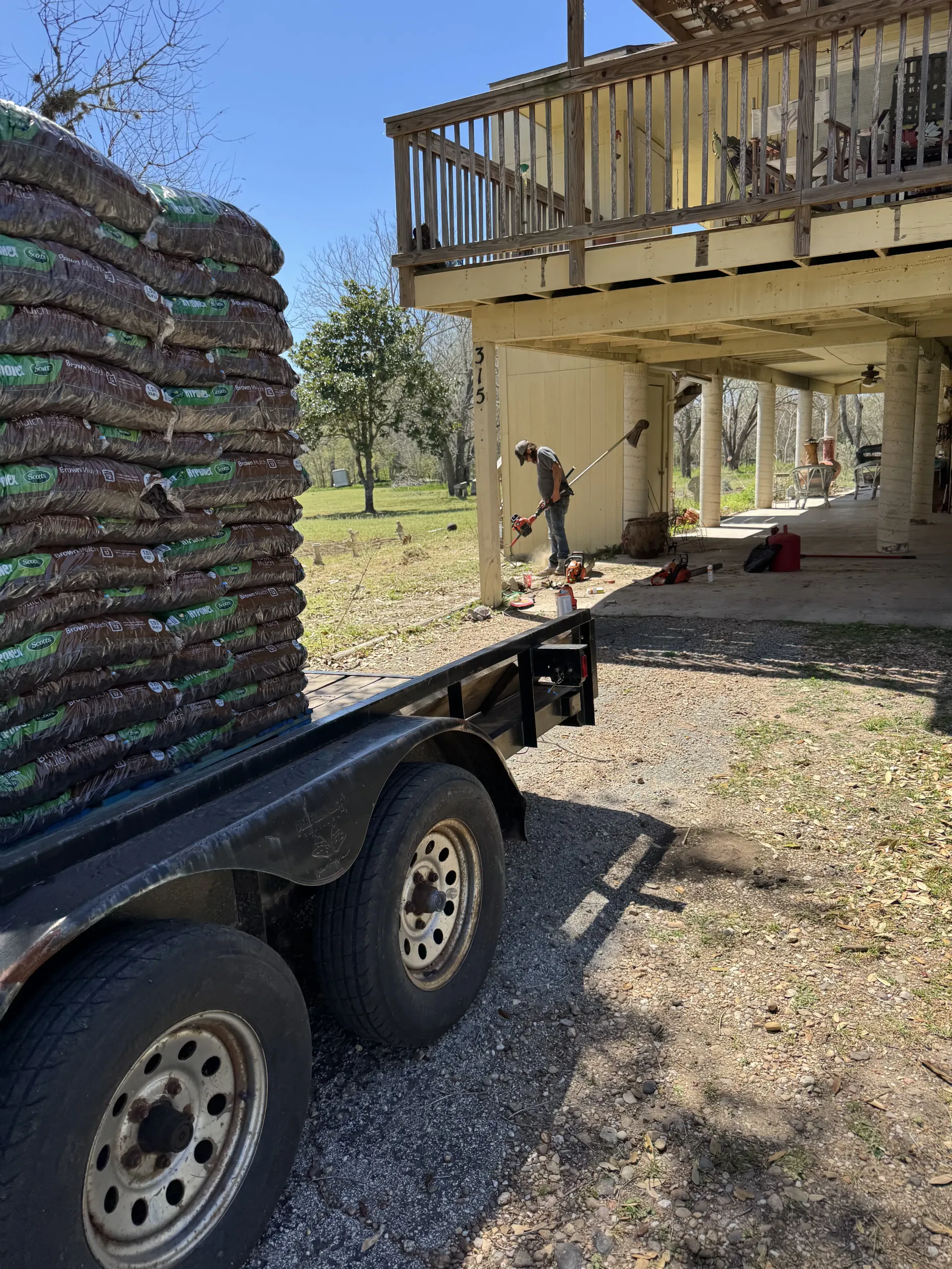 A trailer loaded with stacked bags of mulch sits near a house with a deck while a person works in the yard.