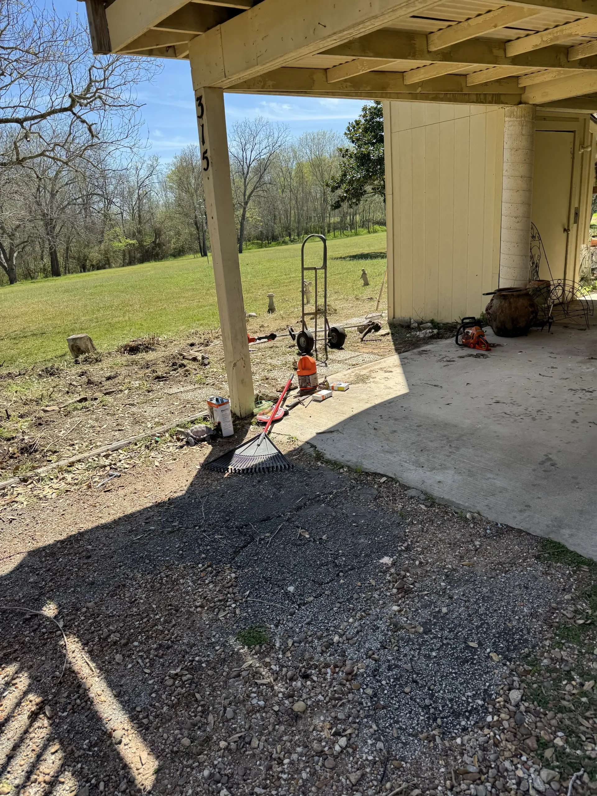 A shaded patio area with a concrete slab, gravel, a yellow support post, and a metal utility cart in the background.