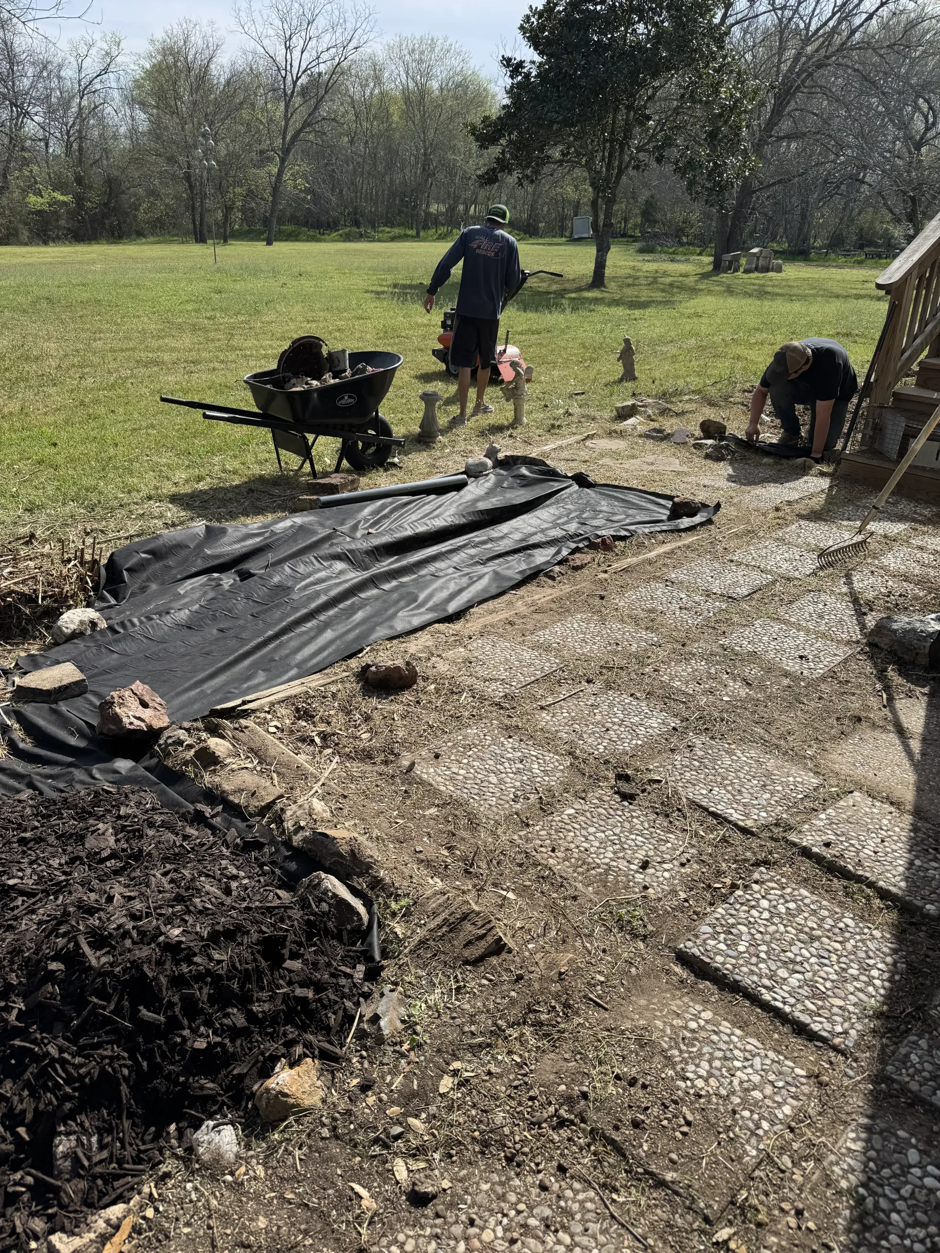 Two people work in a grassy yard, one using a wheelbarrow and the other kneeling near a section of black landscape fabric.