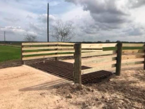 A cattle guard made of dark metal bars set into a dirt path, flanked by wooden fence rails on a rural, overcast day.