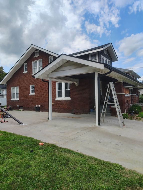 Brick house with white trim and a covered carport, concrete driveway, and a ladder under a partly cloudy sky.