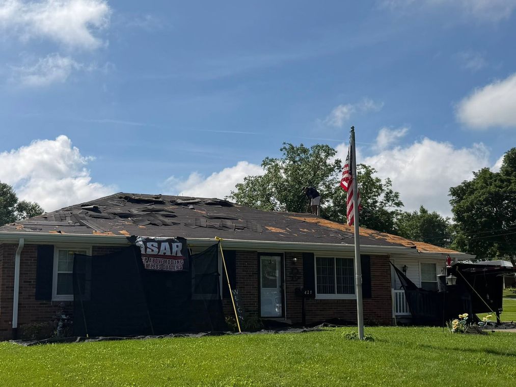 Single-story house with storm-damaged roof, an American flag, and a ladder in the yard under a blue sky.