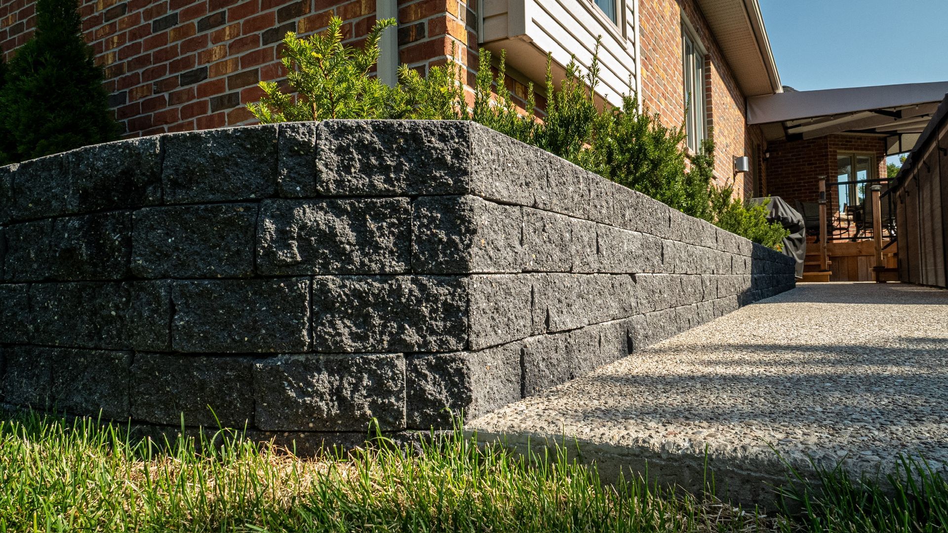 Stone retaining wall beside a brick house, bordering a gravel walkway and grass in sunlight