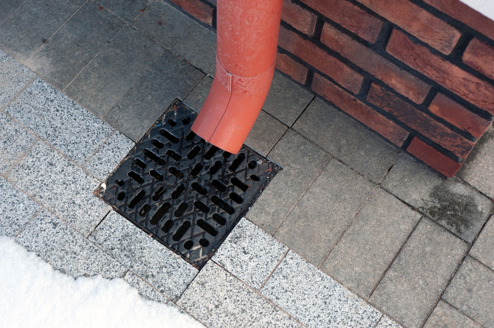 A red pipe is coming out of a drain on a sidewalk next to a brick wall.