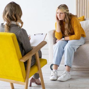 A woman is sitting in a yellow chair talking to another woman.