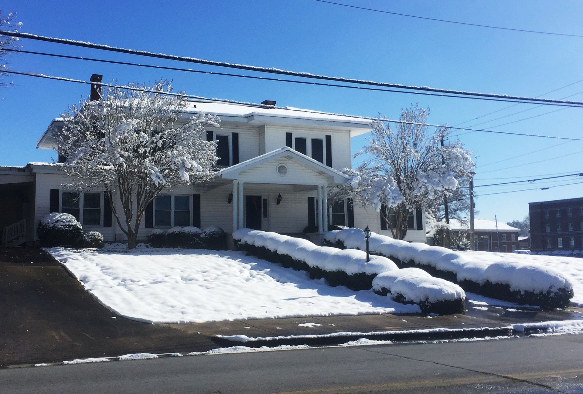 Snow-covered house with a front porch, driveway, and blossoming trees on a sunny day