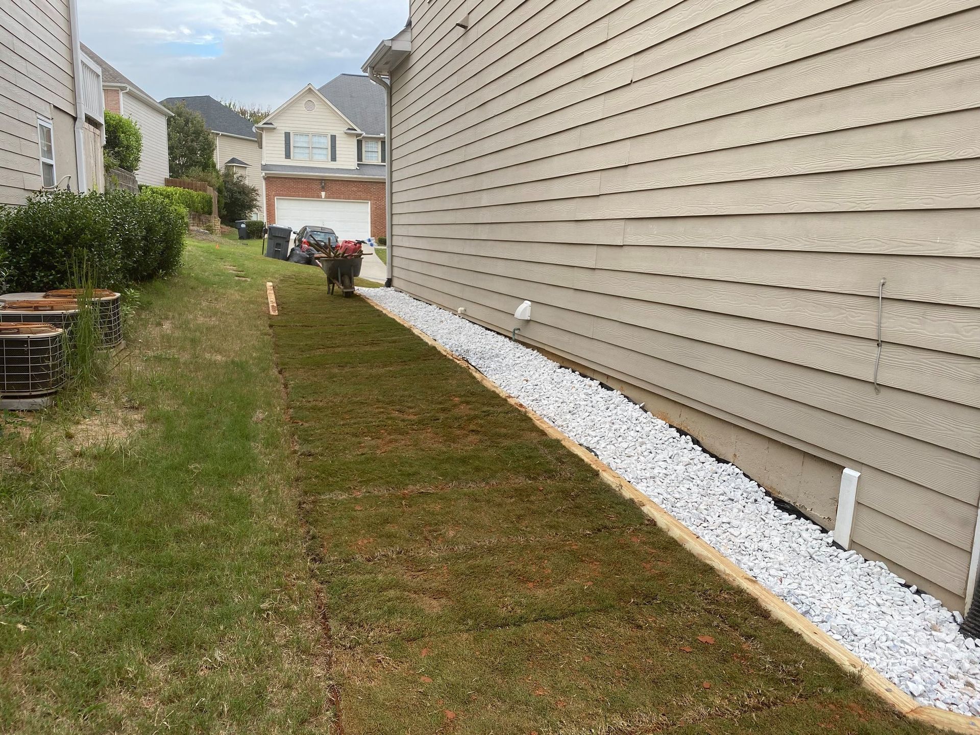 Narrow grass side yard beside beige house, with white stones along the foundation and neighboring homes in view
