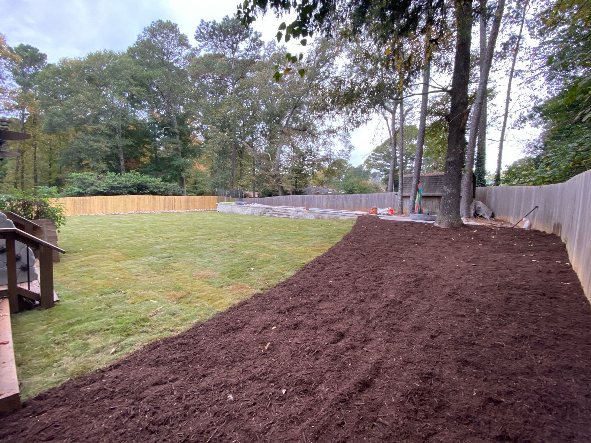 Freshly mulched backyard along a fence, with a grassy lawn and trees in the background