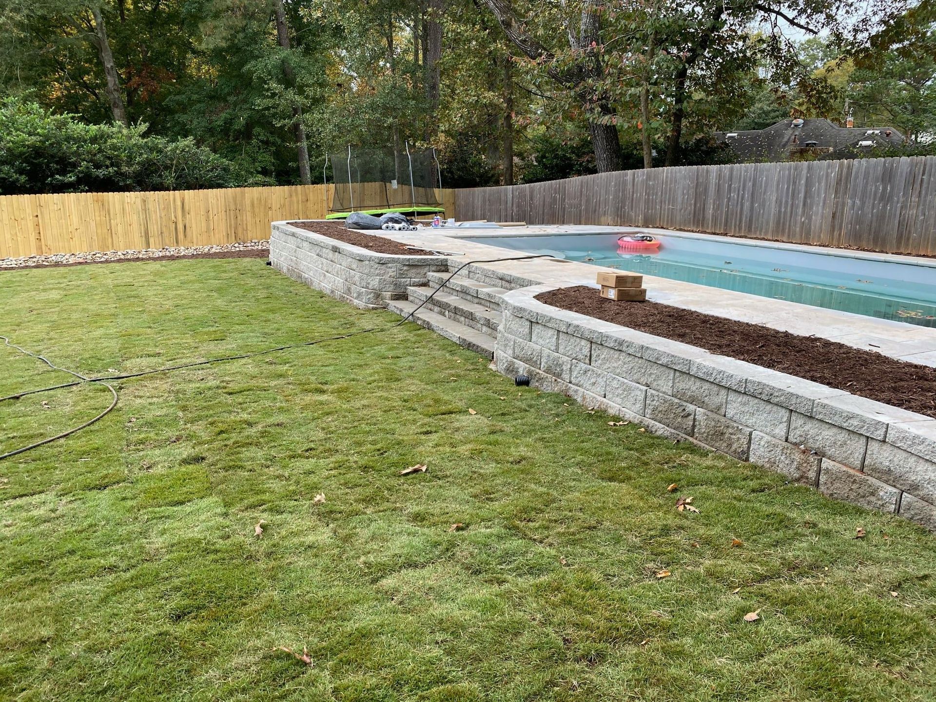 Backyard with grassy lawn, raised pool, and stone retaining wall beside a fenced wooded area